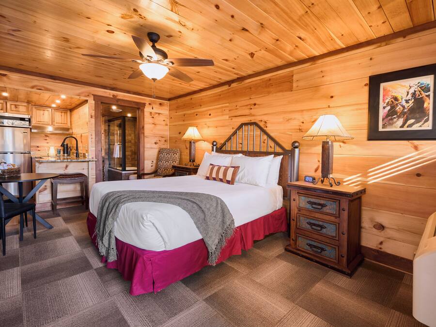 A wooden cabin interior with a bed, bedside lamps, a desk with a chair, and a kitchen area.