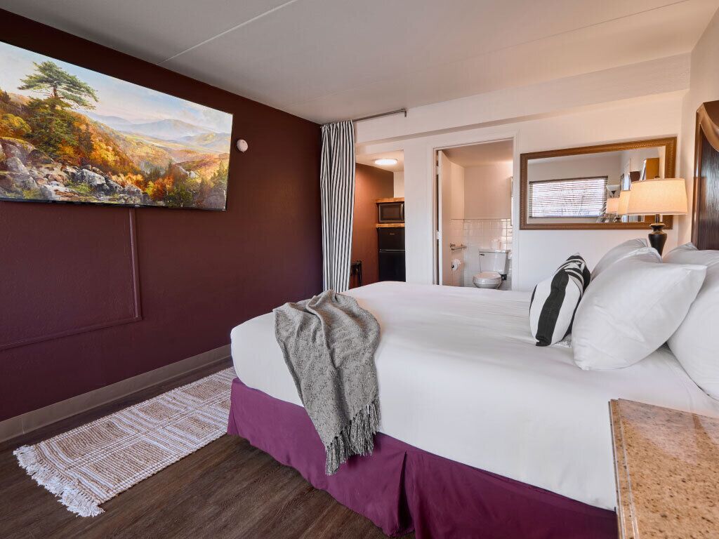 Ein Hotelzimmer mit Bett, einem Wandbild, einem durchgegangene Tür erkennbaren Badezimmer und einem Nachttisch mit Lampe.