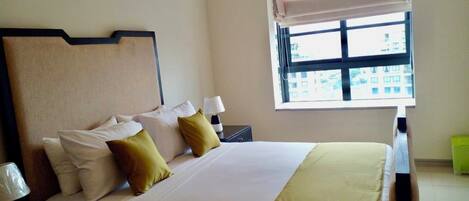 Habitación de hotel con cama, almohadas, una mesita de noche, lámpara y ventana con vista a edificios.