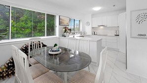 A modern kitchen with white cabinets, a glass dining table, and a view of greenery through the windows. - Relaxing Getaway Steps Beach With Pool (Hervey Bay)