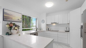 A modern kitchen with white cabinetry, a large island, a window with blinds, a clock, and a coffee maker. - Relaxing Getaway Steps Beach With Pool (Hervey Bay)