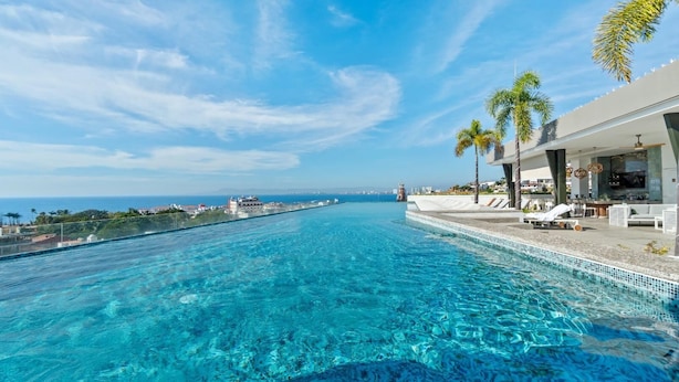 A clear blue infinity pool with a view of the ocean and a modern building with lounge chairs and palm trees. - Rooftop Pool + Hot tub + Gym | Romantic Zone Haven (Puerto Vallarta)