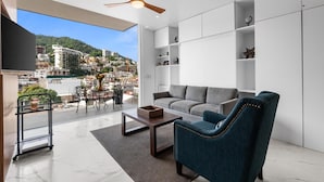 A modern living room with a sofa, armchair, and coffee table, a balcony with a view of a cityscape, and a ceiling fan. - Rooftop Pool + Hot tub + Gym | Romantic Zone Haven (Puerto Vallarta)