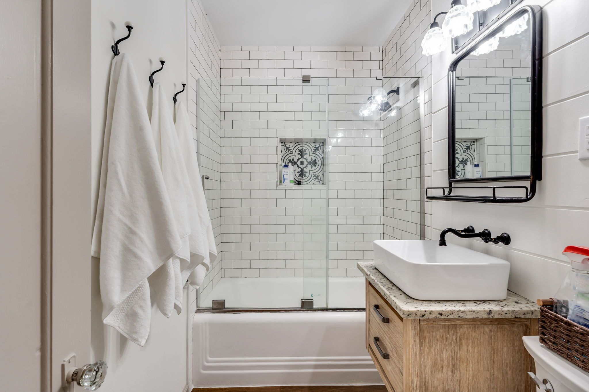 A bathroom with white tiled walls, a white bathtub, a white sink with a black faucet, and a mirror with lights.