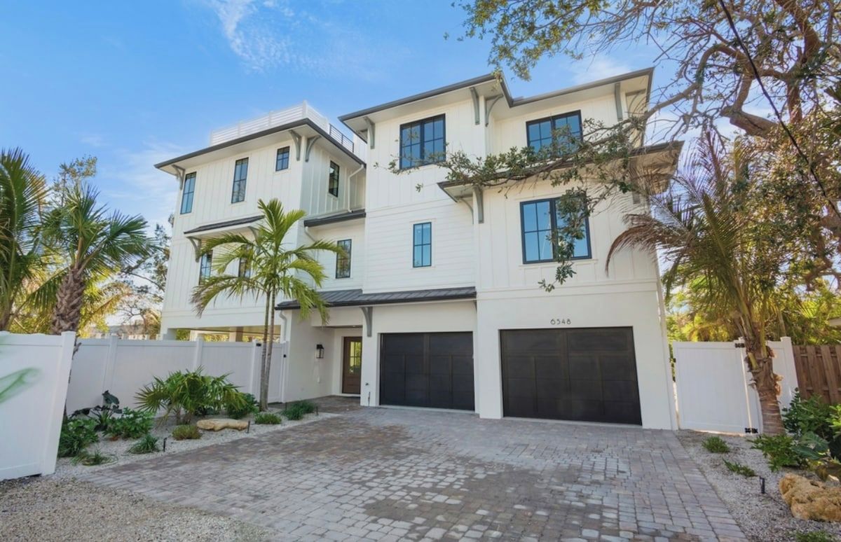 A modern two-story house with a garage, surrounded by a white fence and palm trees.
