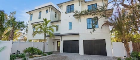 Uma casa moderna de dois andares com garagem, cercada por um muro branco e palmeiras.