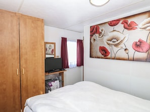 Une chambre à coucher avec un lit, une armoire en bois, un téléviseur posé sur un meuble, un tableau représentant des fleurs et une fenêtre avec des rideaux.