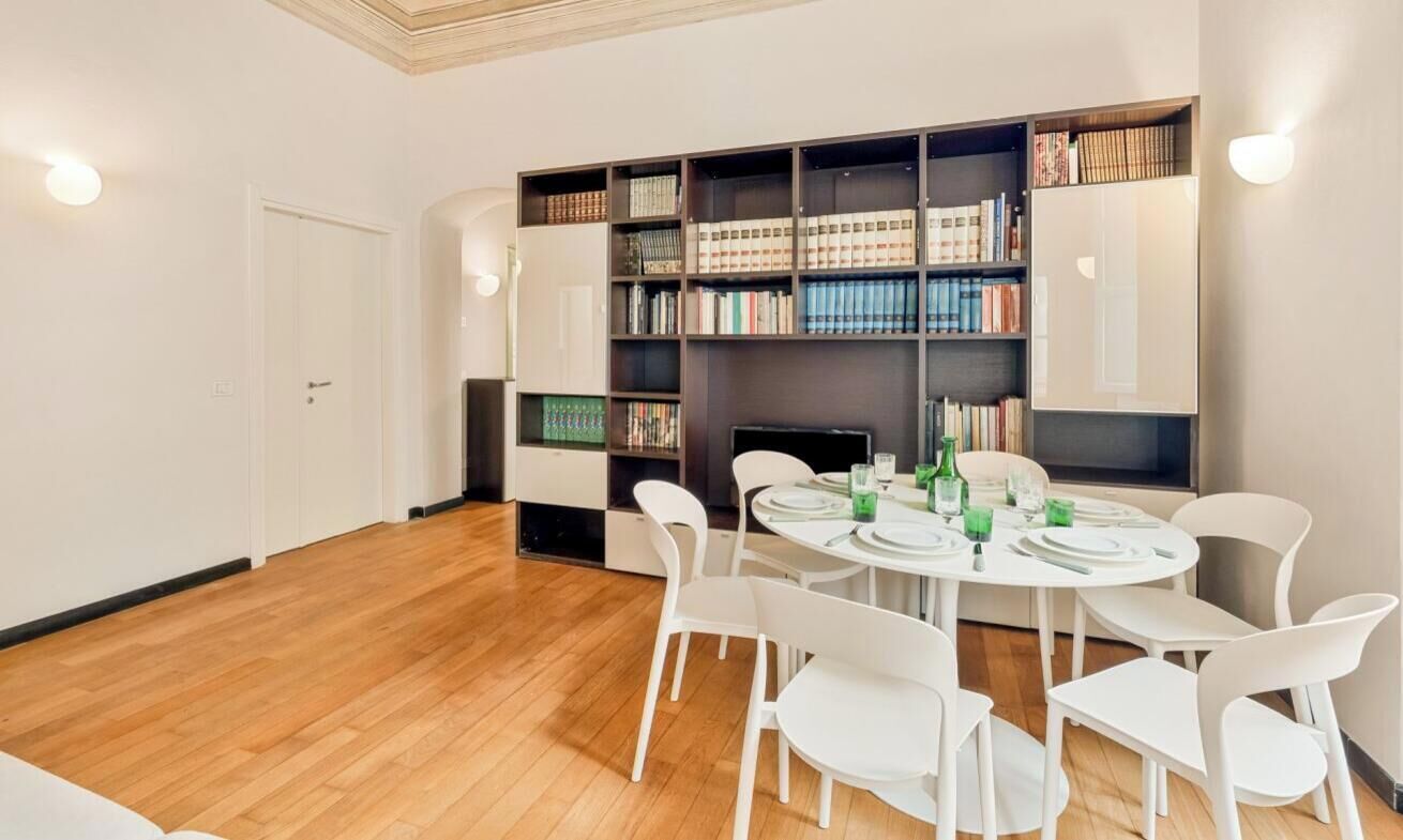 Zona pranzo con un tavolo rotondo apparecchiato per quattro persone, circondato da sedie bianche. Una libreria piena di libri è appoggiata alla parete, e una TV a schermo piatto è montata sopra il tavolo.