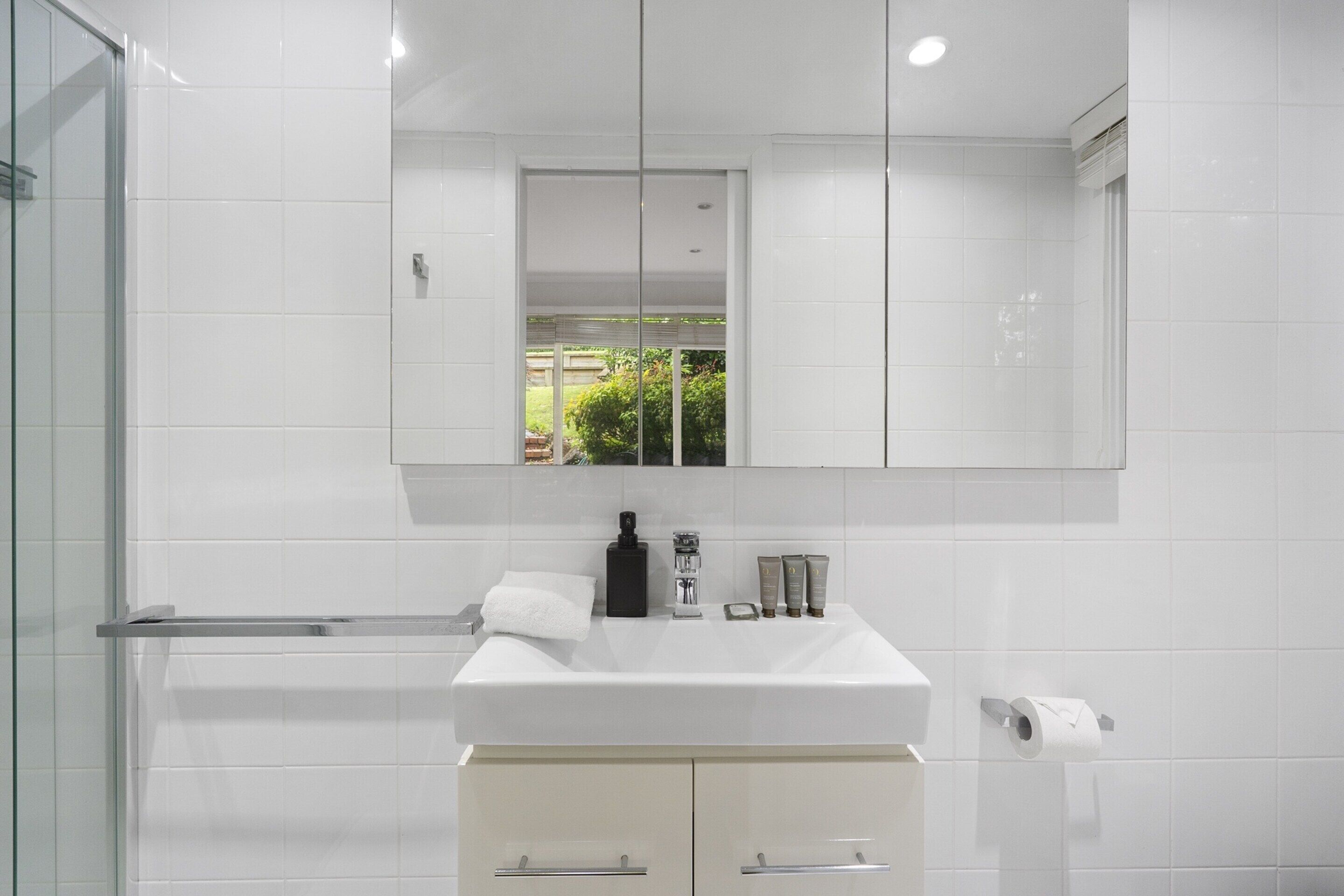 A modern bathroom with a large mirror, white tiles, a glass shower enclosure, and a white sink with toiletries.