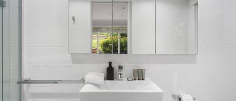 A modern bathroom with a large mirror, white tiles, a glass shower enclosure, and a white sink with toiletries.