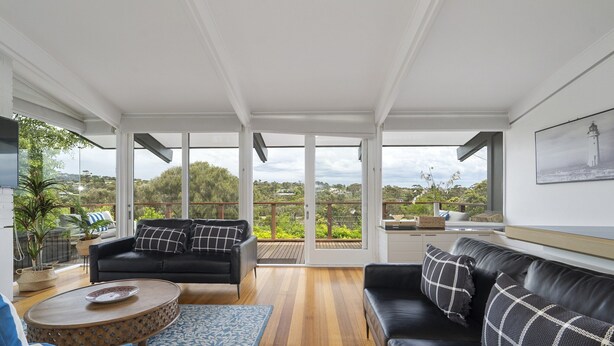 Ein modernes Wohnzimmer mit einer schwarzen Ledersofa, einem Holztisch und einem Teppich mit blauem Muster. Große Fenster bieten einen Ausblick ins Freie.