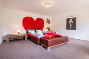 Une chambre à coucher avec une grande tête de lit en forme de cœur, un lit avec une literie rouge et bleue, une table de chevet en bois et une œuvre d’art accrochée au mur.