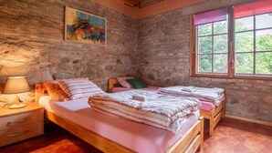 Een slaapkamer met een stenen muur, een bed met roze beddengoed, een nachtkastje met een lamp en een raam met uitzicht op groen.