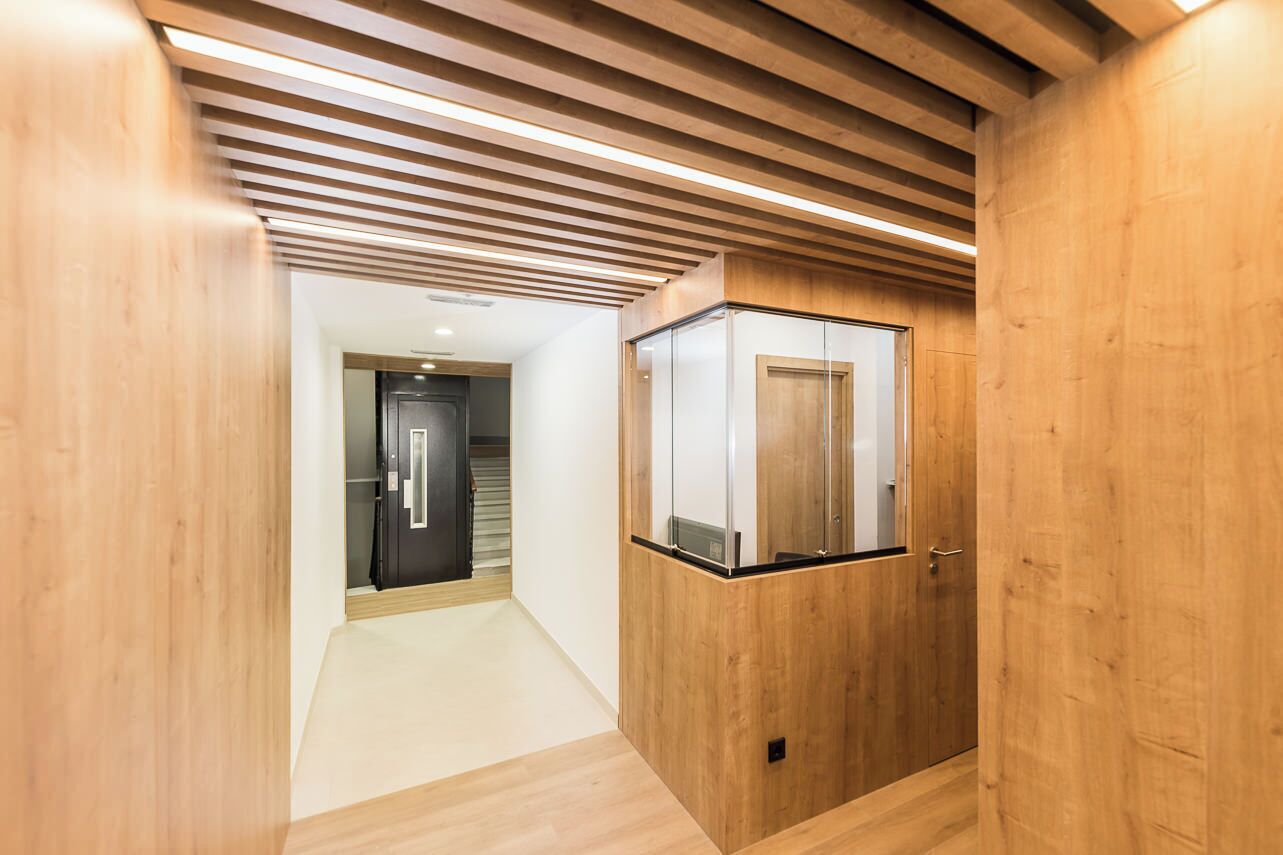 Een hal met houten wanden en plafond, een glazen lift en een deur naar een andere kamer.