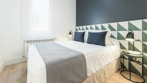 Een net opgemaakt bed met een wit-grijs beddegoed, een donkere muur met geometrische patronen en een klein nachtkastje met een lamp.