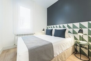 Una cama bien hecha con una colcha blanca y gris estampada, una pared oscura con diseños geométricos y una mesita de noche con una lámpara.