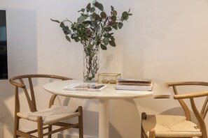 Una mesa redonda blanca con dos sillas de madera, un jarrón con follaje verde y una pila de libros.