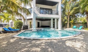 Un área de piscina moderna al aire libre con una zona de estar techada, palmeras y un edificio de dos pisos.