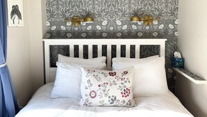 Un dormitorio con cama, ropa de cama blanca, un cojín floral, una pared con estampado de papel tapiz floral y dos lámparas fijadas a la pared.