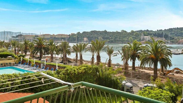 Ein Poolbereich mit Liegestühlen, Palmen und Blick auf Wasser und Gebäude im Hintergrund.