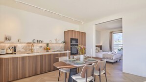 Een moderne keuken met een centraal kookeiland, een eethoek met stoelen, en uitzicht op een woonkamer met een bank en een bed.