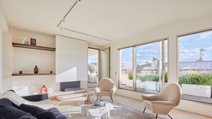 Een moderne woonkamer met een bank, twee fauteuils, een salontafel en een televisie. Grote ramen bieden uitzicht op de stad.