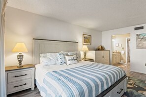 A bedroom with a bed, two bedside tables, a nightstand, a dresser, and a door leading to another room. - Edgewater Villas 1BR | Pools, Beach Access, & Golf (Panama City Beach)