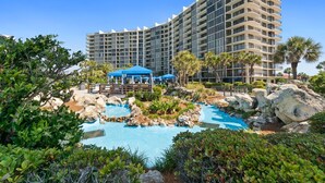 A hotel with a large pool surrounded by rocks and greenery, a blue canopy, and a modern building with multiple balconies. - Edgewater Villas 1BR | Pools, Beach Access, & Golf (Panama City Beach)