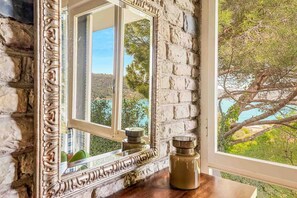 Une pièce avec un mur en pierre, un grand miroir décoratif et une vue sur un arbre et des collines à l’extérieur.