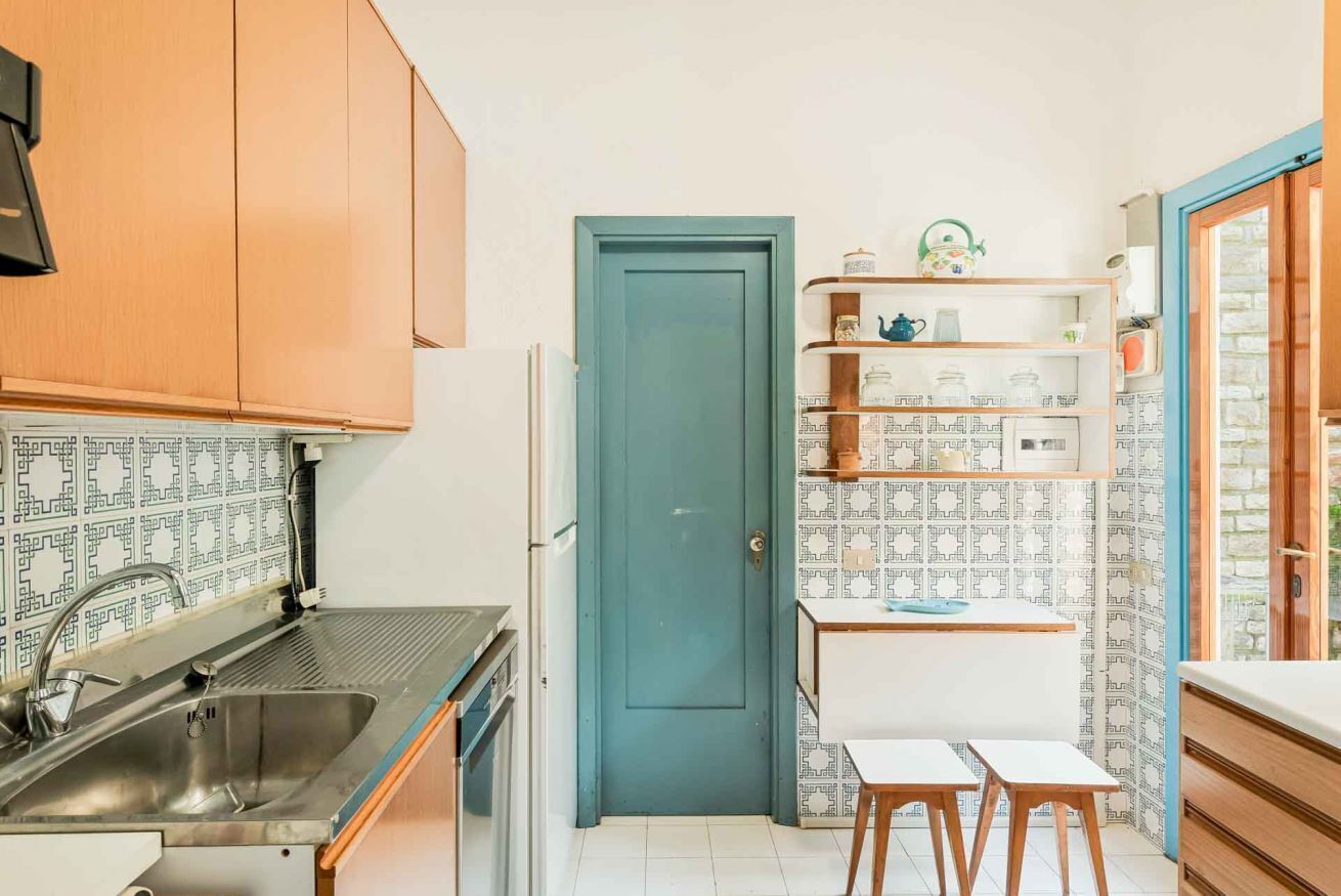Een kleine keuken met houten kastjes, een blauwe deur en een eethoek met krukjes.