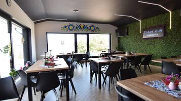 A restaurant with multiple tables and chairs, a green wall with a sign, and large windows allowing natural light.