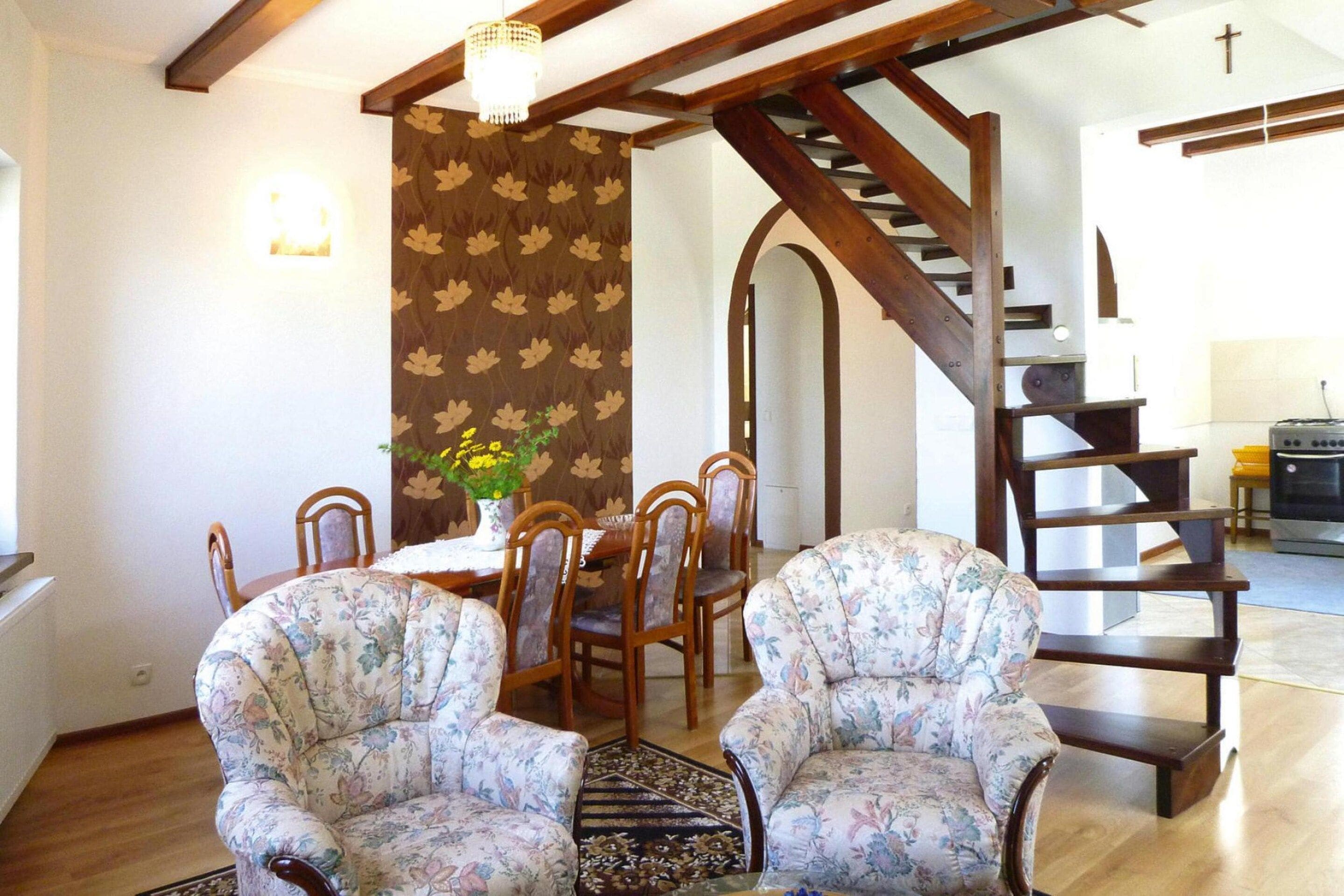 A dining area with floral-patterned chairs, a wooden table, and a vase with yellow flowers. A staircase with wooden railings leads to another room.