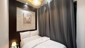 A neatly made bed with white linens and a rolled towel, a small bedside table with a lamp and a plant, and a large window with grey curtains. - Attraction Partition Room in Tecom (Dubai)