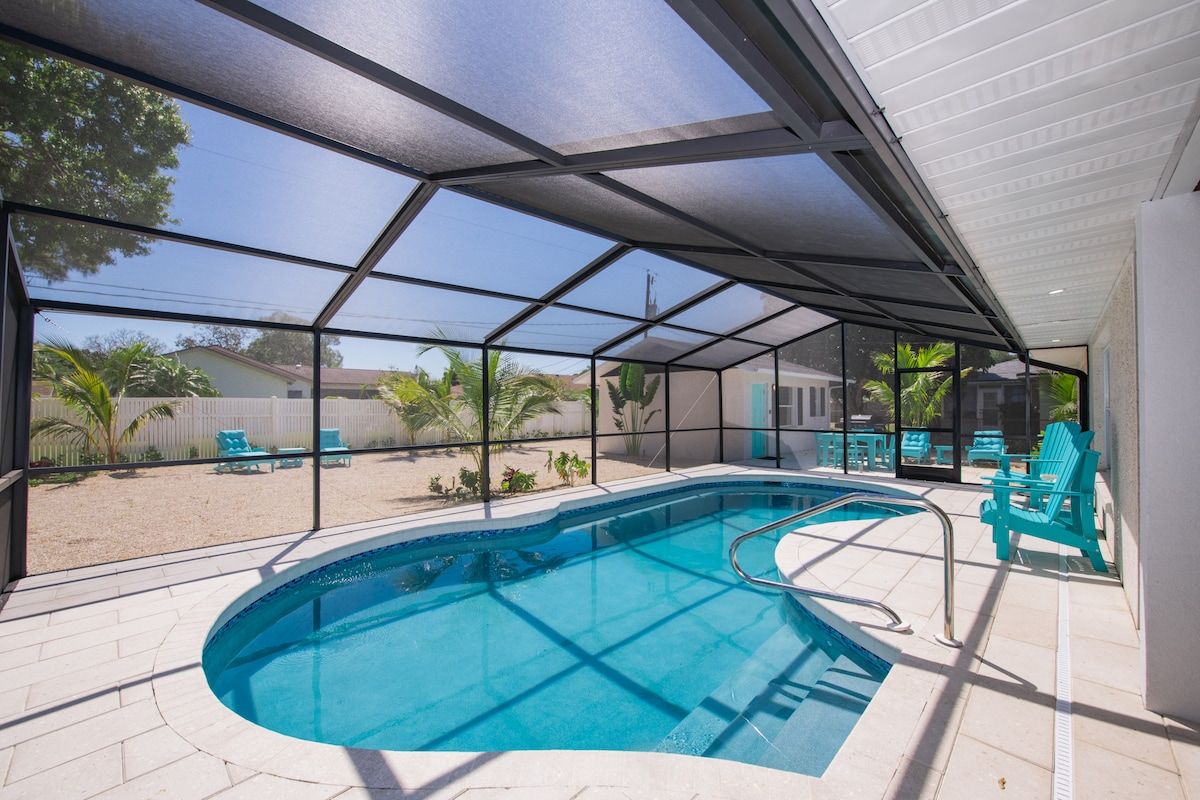 An indoor swimming pool with a glass enclosure, surrounded by lounge chairs and a view of the outdoors.
