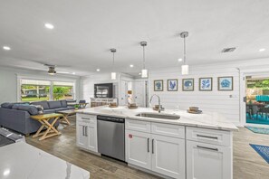 A modern kitchen with white cabinetry, a dishwasher, and a large island with a sink. There is a dining area with a gray sofa and a wooden coffee table.