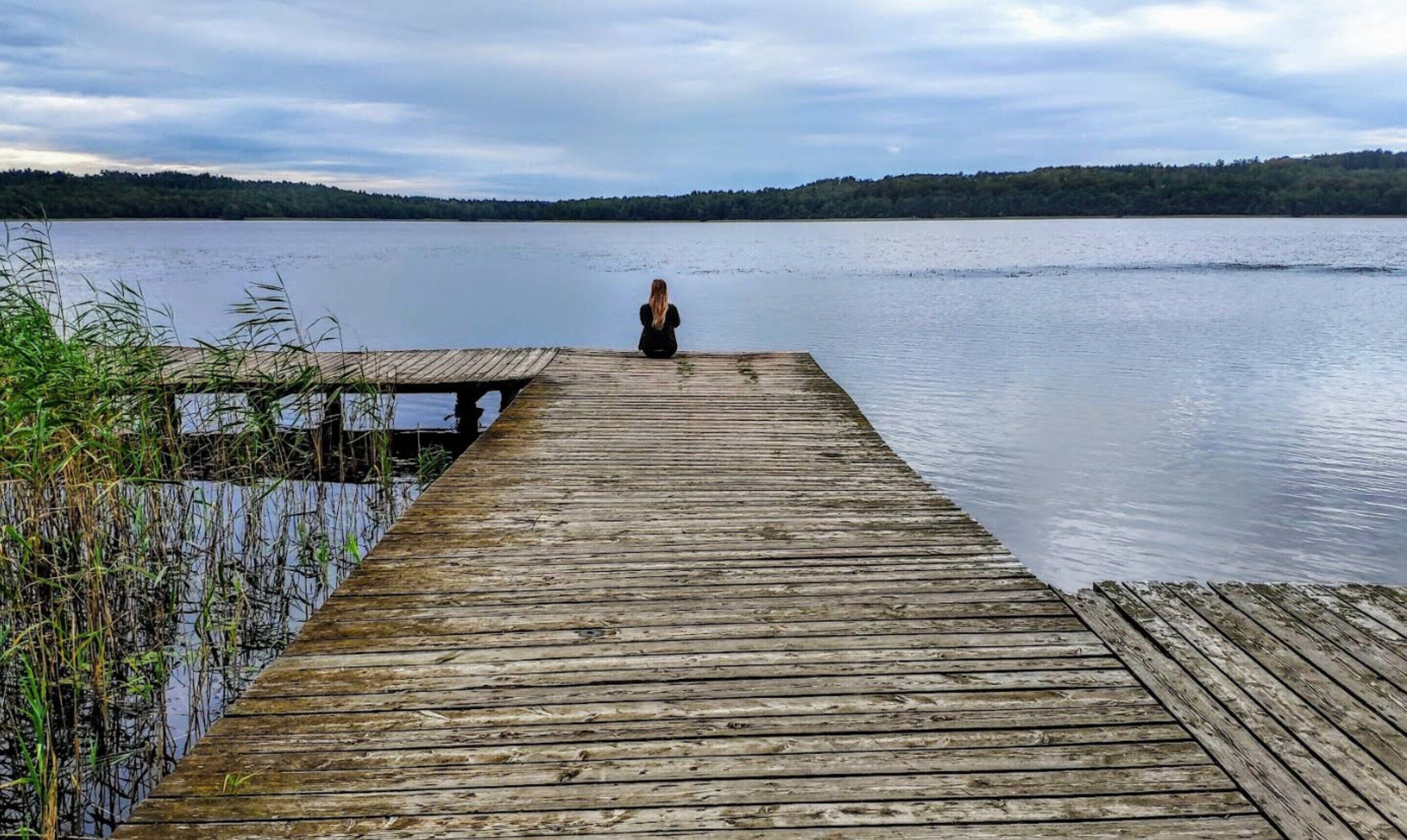A wooden pier extending into a calm lake with a person sitting at the end, surrounded by dense greenery.