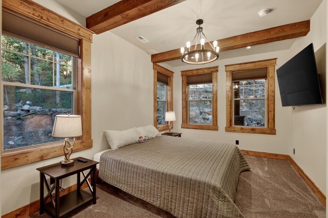Un dormitorio con cama, lámparas de noche, televisor y ventanas con vistas a árboles y rocas.