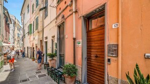 Eine enge Straße mit Häusern an beiden Seiten, einige mit heruntergelassenen Fensterläden. Auf dem Bürgersteig stehen Kübelpflanzen und es gibt Außenplätze.