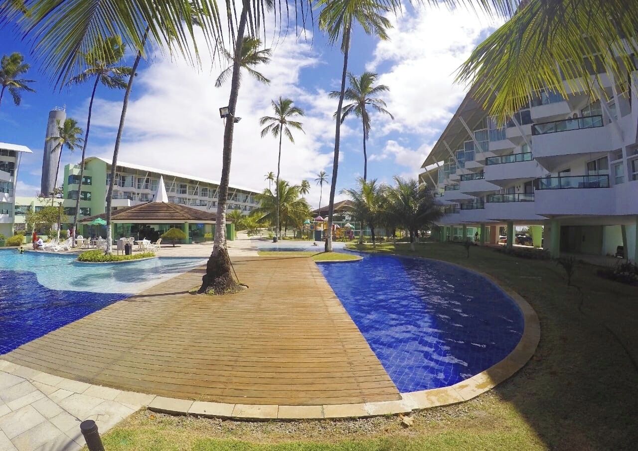 Ein Poolbereich eines Resorts mit Holzterrasse, Palmen und modernen Gebäuden im Hintergrund.