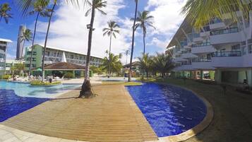 Ein Poolbereich eines Resorts mit Holzterrasse, Palmen und modernen Gebäuden im Hintergrund.