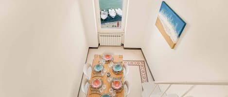 A dining area with a table set for six, a view of boats through a window, and a painting on the wall.