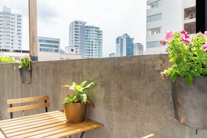 Un balcón con una mesa de madera, dos plantas en macetas y vistas a edificios de la ciudad.