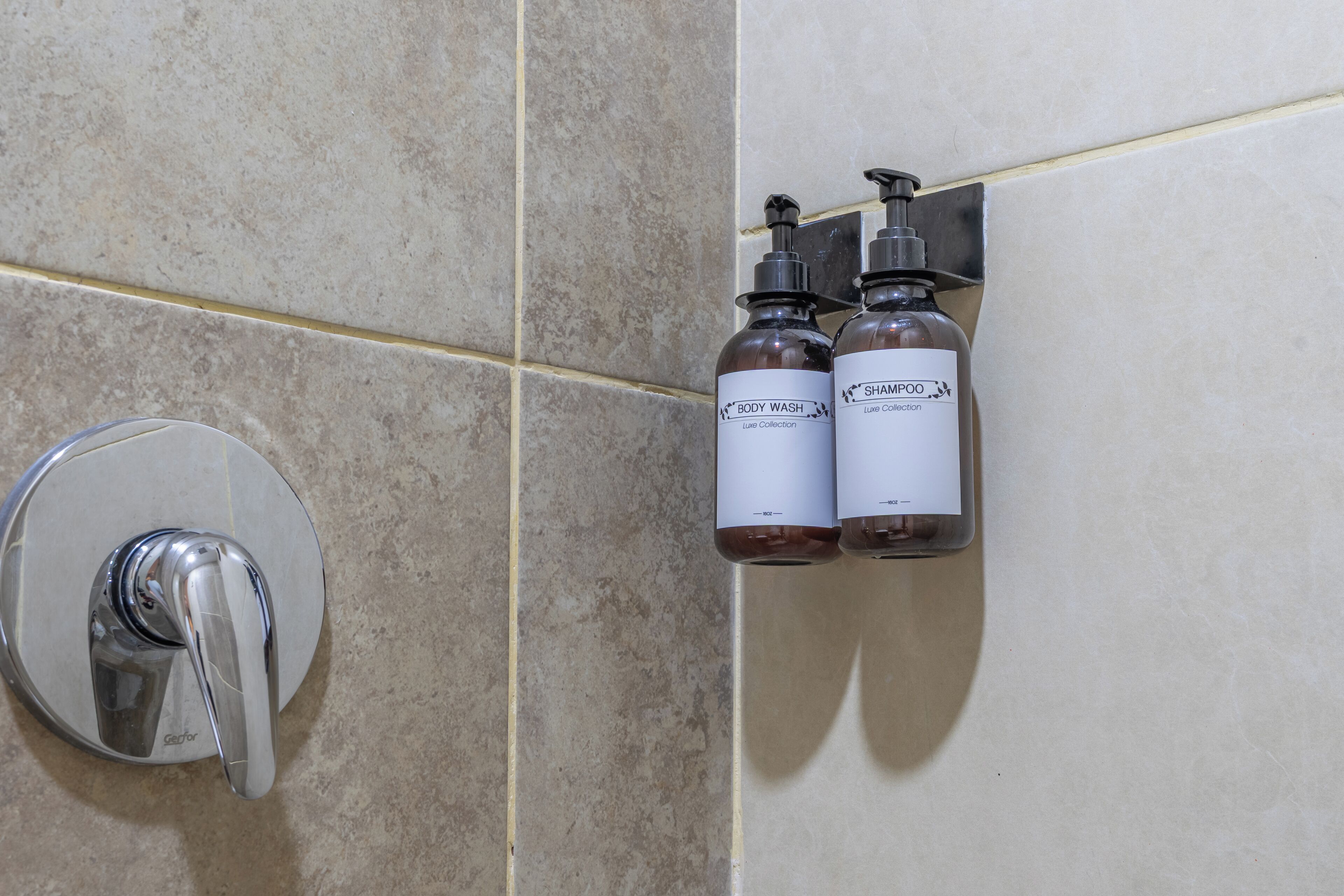 A bathroom with a showerhead, two body wash bottles, and a soap dispenser on a wall.