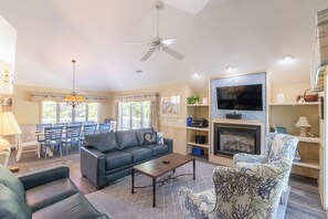 A living room with a sofa, armchair, coffee table, fireplace, TV, and dining area with chairs and a table. - Sun Realty pet-friendly oceanside Duck home with hot tub (Duck)