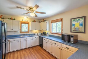 A kitchen with white cabinets, a microwave, a ceiling fan, and a window with a view of trees. - Sun Realty pet-friendly oceanside home with private pool! (Duck)