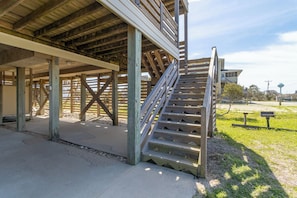 A wooden structure with a staircase leading up to a covered area, surrounded by a grassy area with a bench and a sign. - Oceanfront home w/ private beach walkway, ocean views, & hot tub! (South Nags Head)