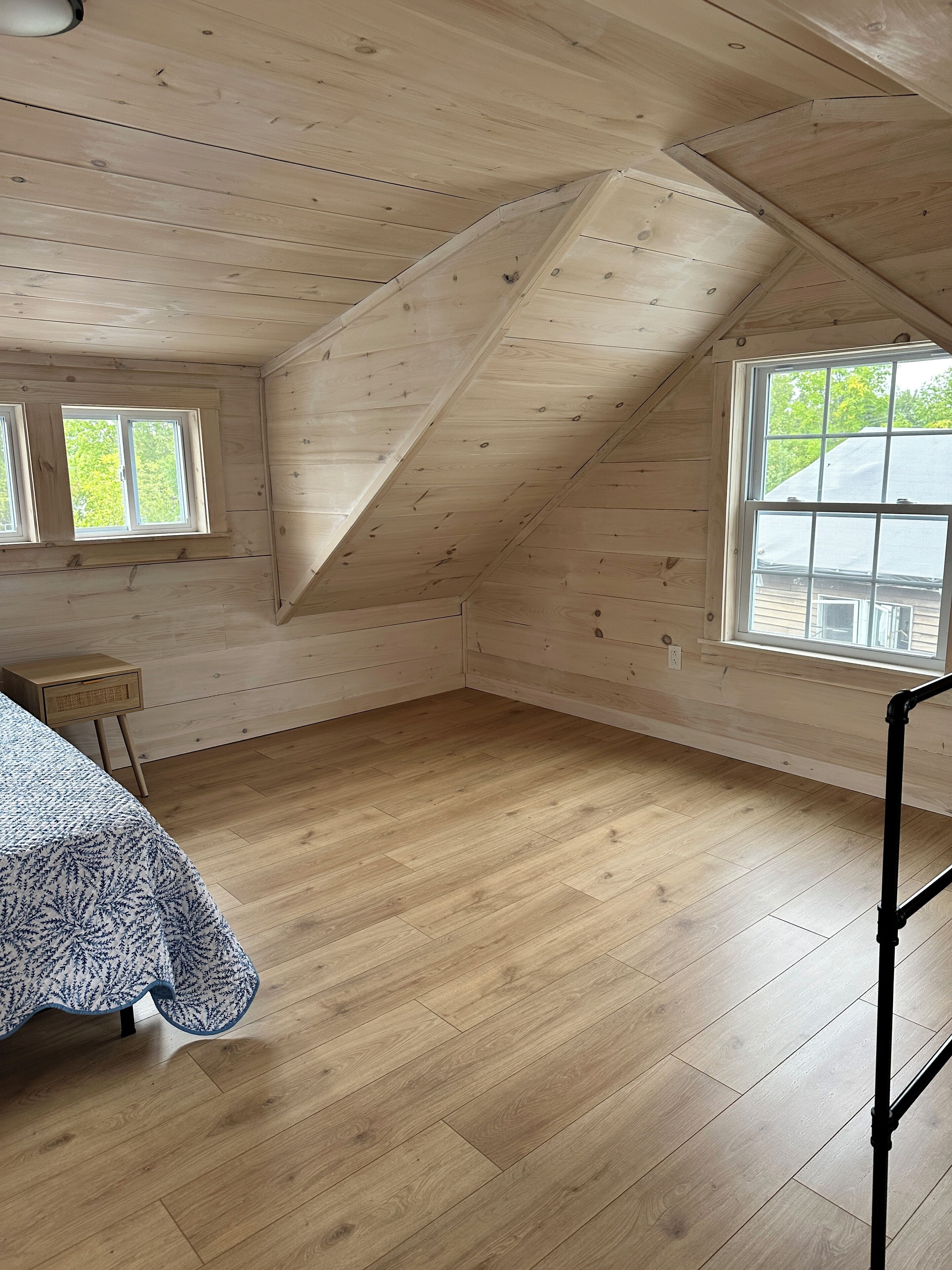 A room with wooden walls and floors, a sloped ceiling, two windows, and a bed with a patterned bedspread.