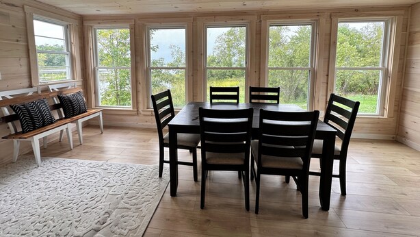 Un comedor con mesa y sillas de madera, ventanas amplias con vista a follaje, un banco con cojines y una alfombra estampada.