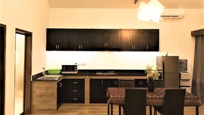 A modern kitchen with dark cabinets, a dining table with chairs, and a hanging light fixture. - Umalas Studio Near Canggu & Seminyak • Fast Wi-Fi + Kitchen (Badung Regency)