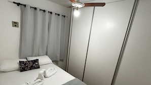 Ein ordentlich bezogenes Bett mit grauer Decke, einem Kissen und einem Kissen mit Blumenmuster. Über dem Bett befindet sich ein Deckenventilator, und an der Decke eine Leuchte.